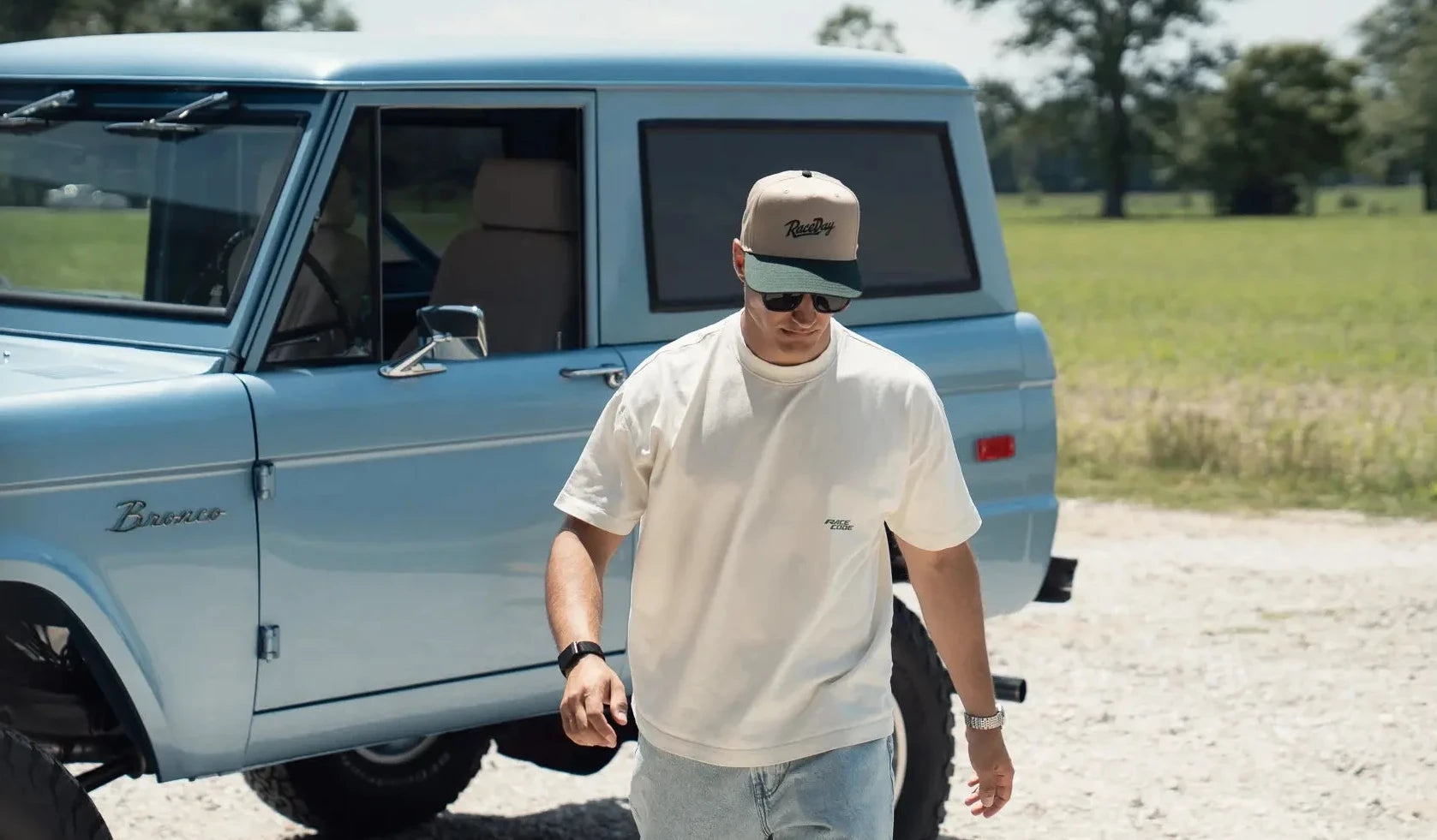 A man wearing a beige cap, white t-shirt, and light blue jeans walks in front of a light blue vintage pickup truck on a gravel path with a green field in the background.