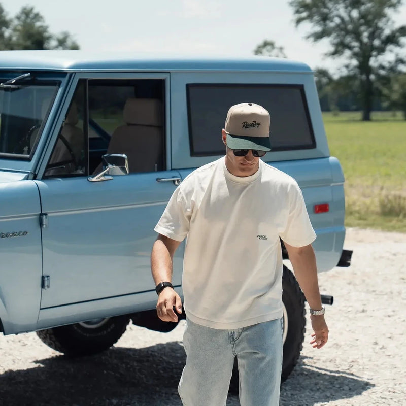 A man wearing a Race Day, Race Code t-shirt, and light blue jeans walks in front of a 1971 Ford Bronco truck on a gravel path with a green field in the background.