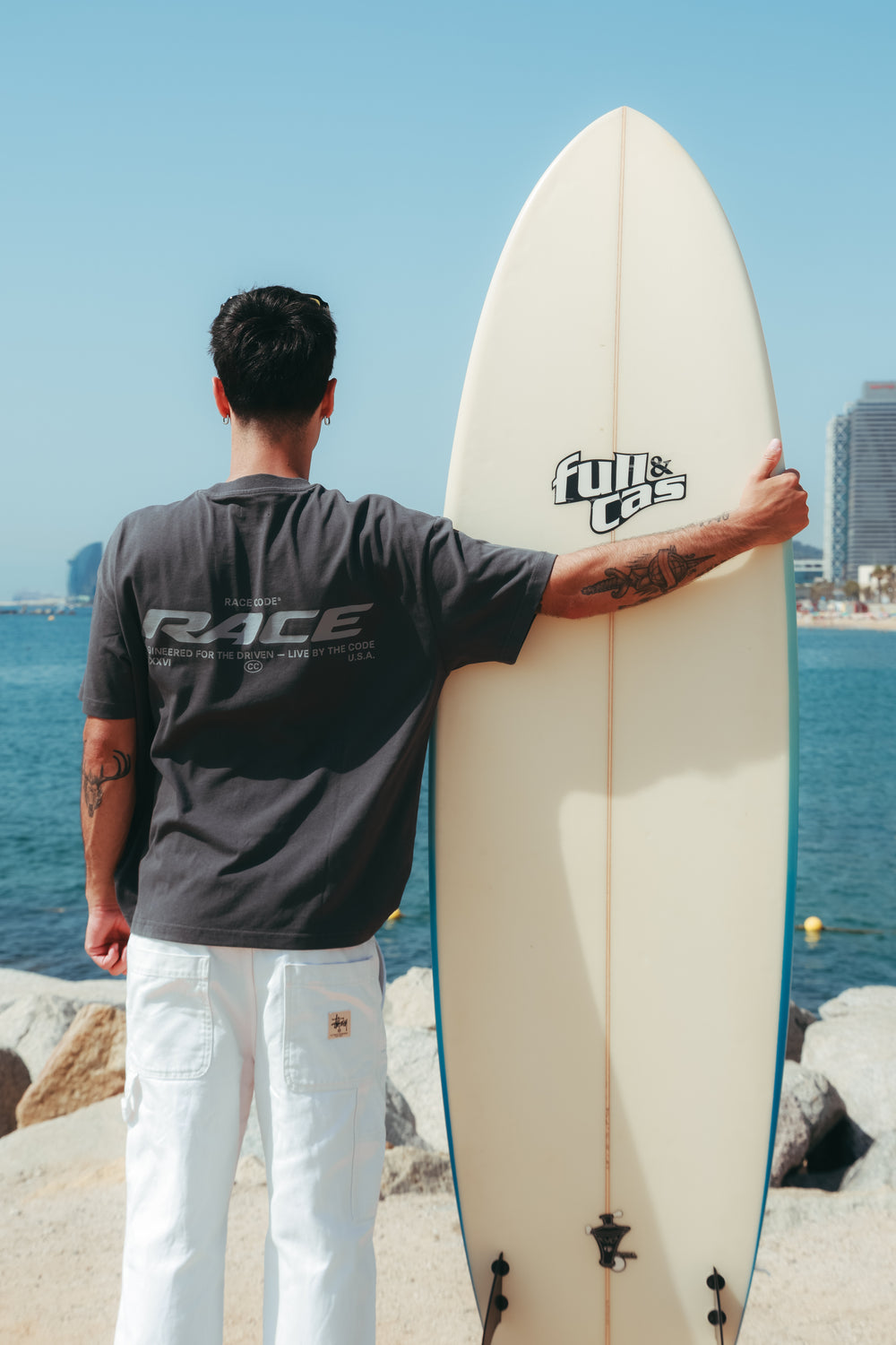 Race Tee Dark Grey con una tabla de surf en Barcelona