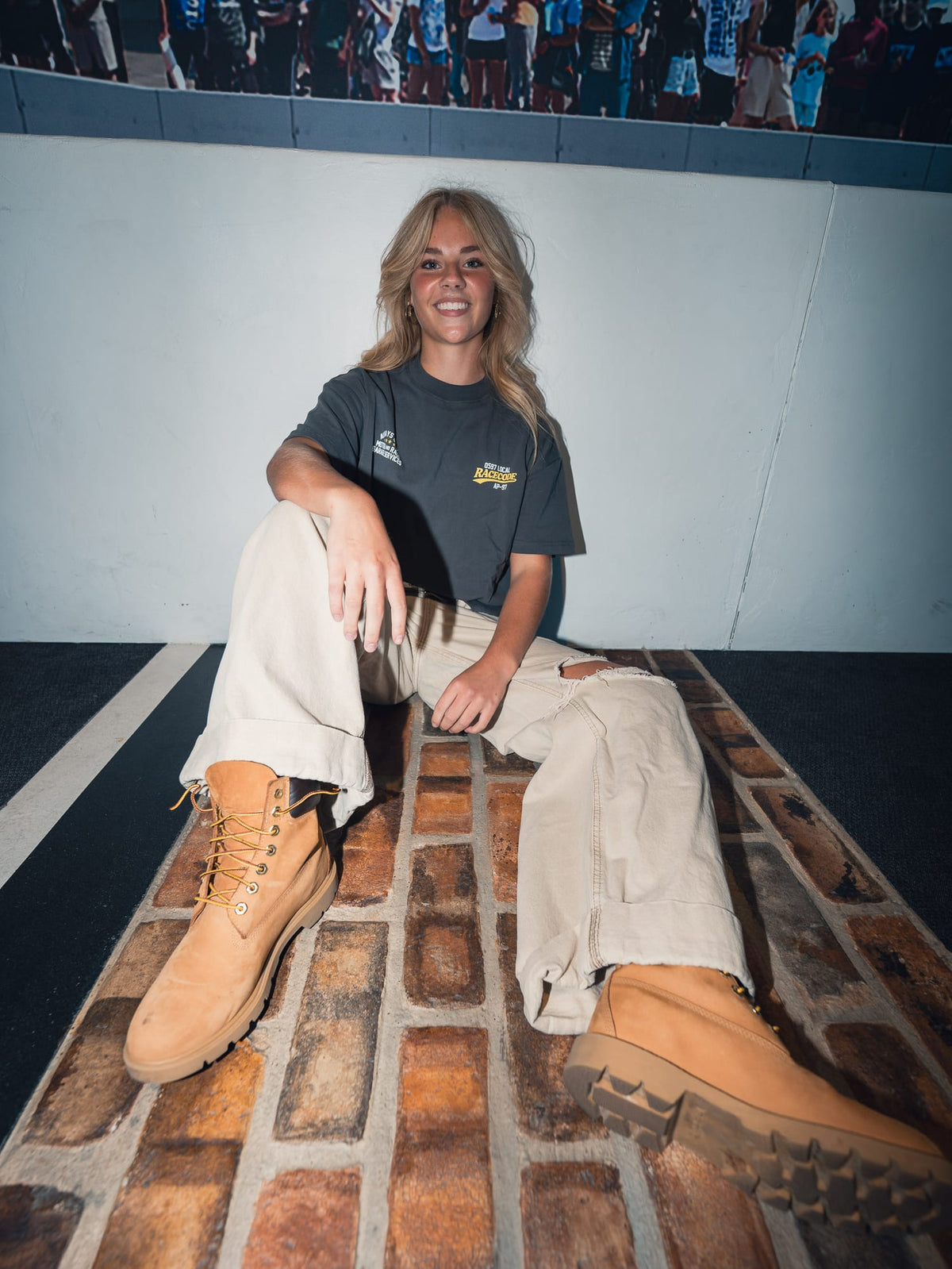 Model sitting on a brick floor wearing the Garage Tee Dark Grey